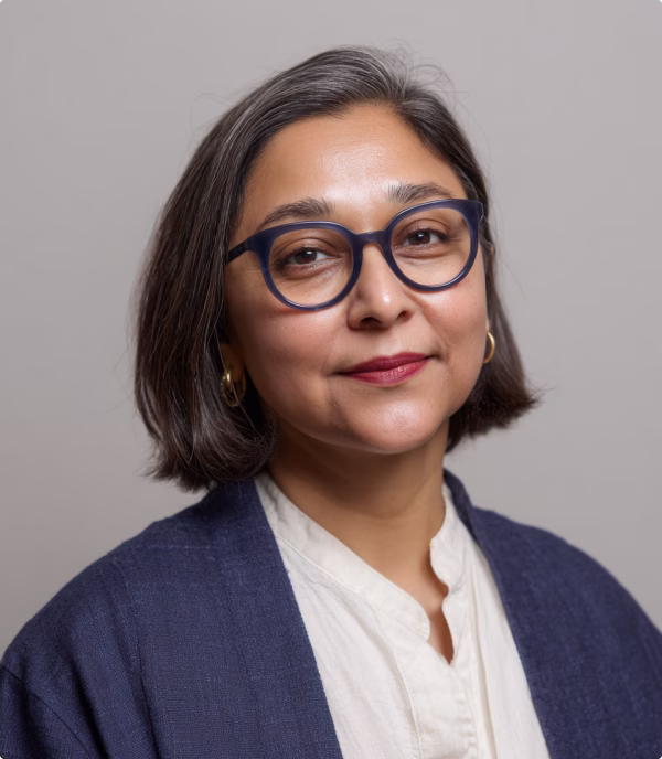 Portrait of a middle-aged woman with short dark hair, wearing blue glasses, gold hoop earrings, a white blouse, and a dark blue cardigan.